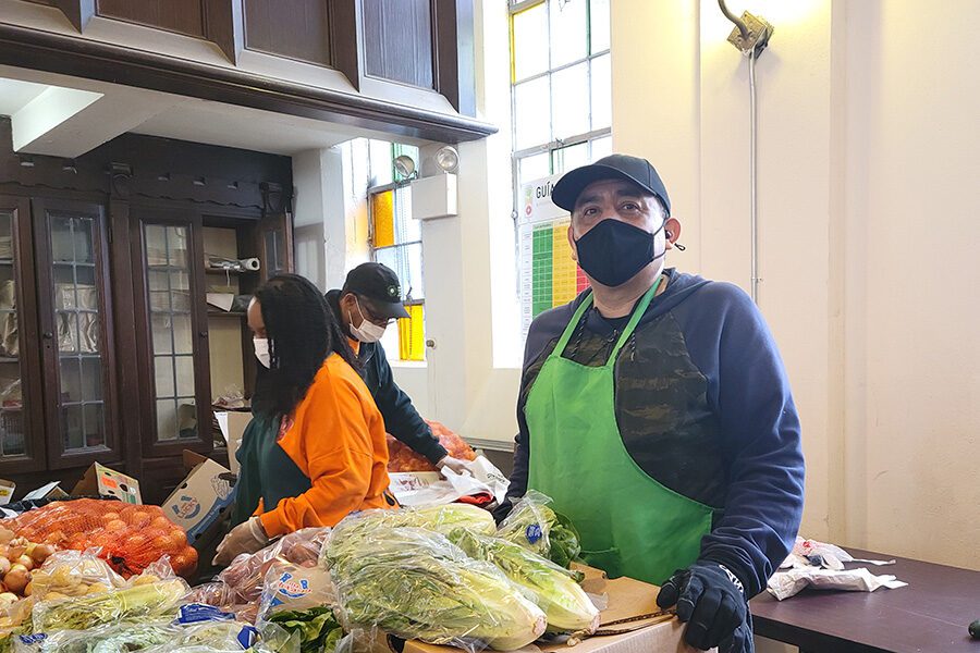a man in a mask and apron looks at the camer from behind a produce table at a food pantry