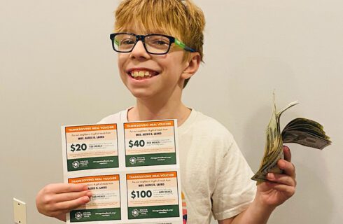 a boy in glasses holds a mailing and some money while smiling at the camera