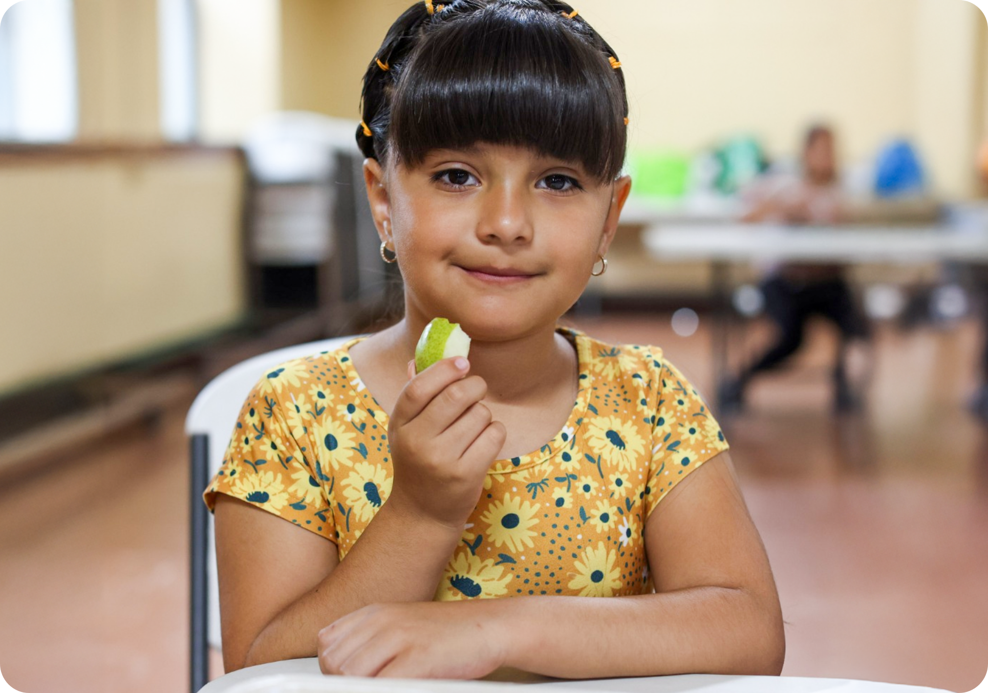 A young girl in the foreground smiles holding an apple while another child and adult are shown in cafeteria setting behind her.