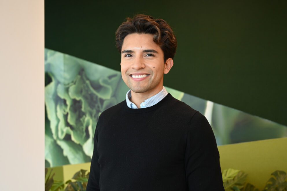 Adult male with styled hair and a black sweater, smiling at the camera Infront of a green, textured wall.