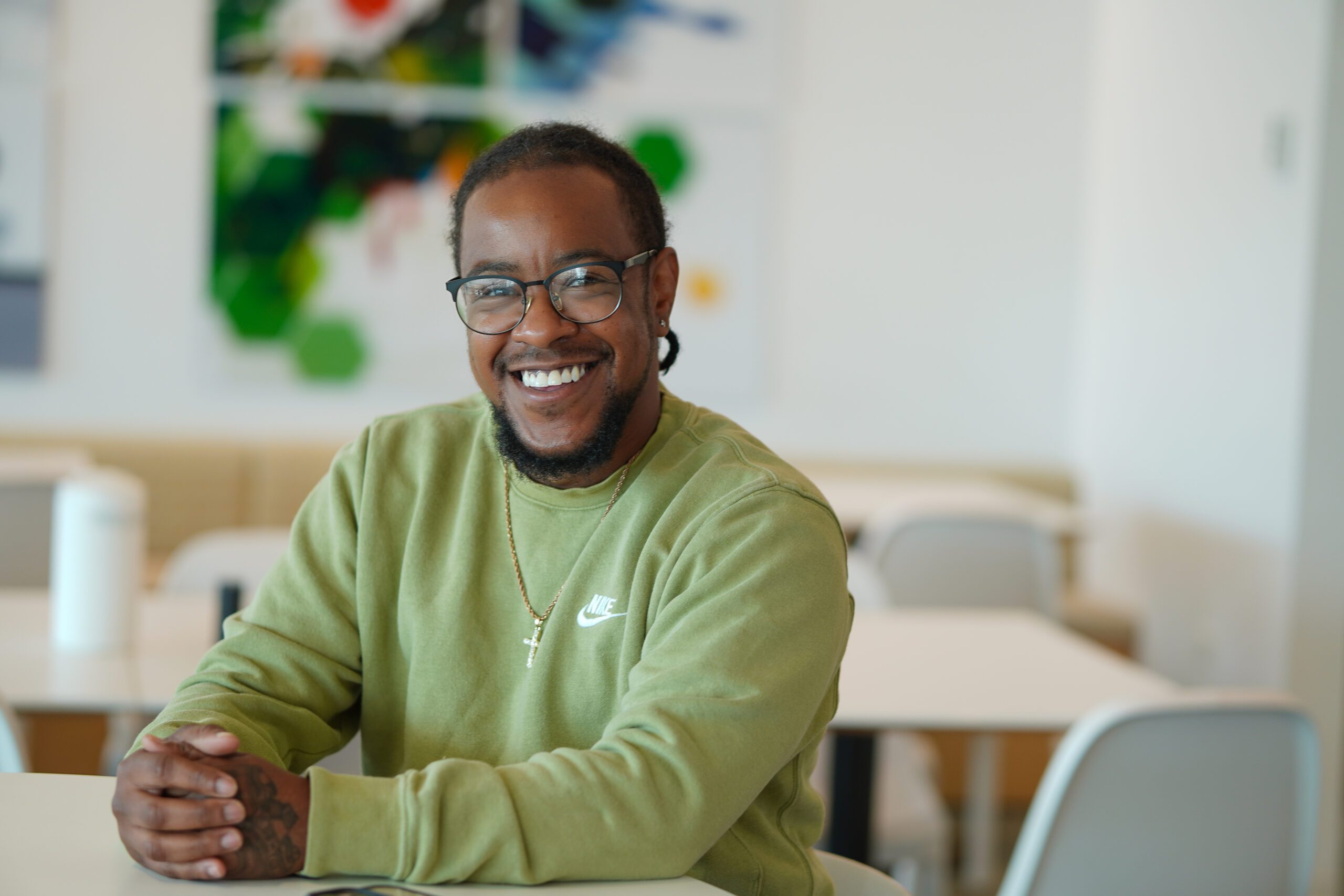 Man wearing a green Nike sweatshirt and glasses, sits in the distance while making eye contact with the camera and smiling.